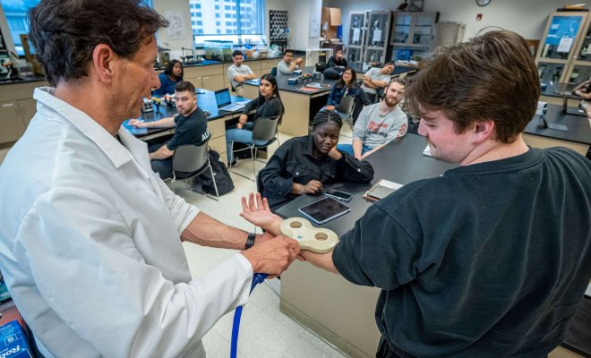 A teacher holds a magnetic coil designed to detect neural activity over a student's arm in front of a lab full of students.