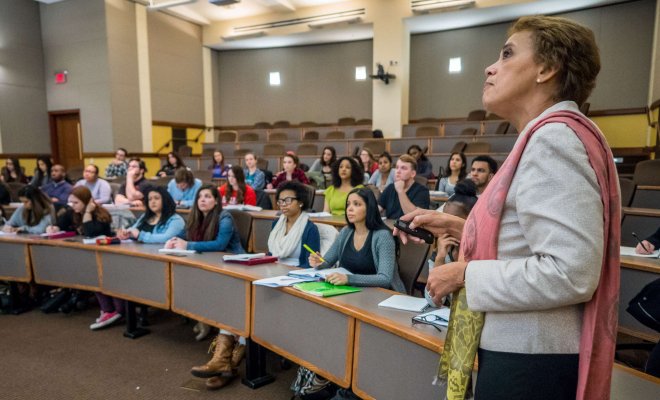 A college professor stands at the front of a lecture hall, addressing a classroom filled with students who are seated in curved rows and taking notes.