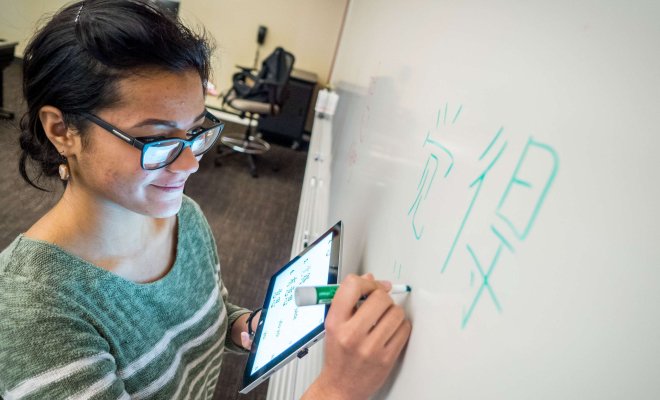 A student stands at a whiteboard in a classroom, writing Chinese characters.
