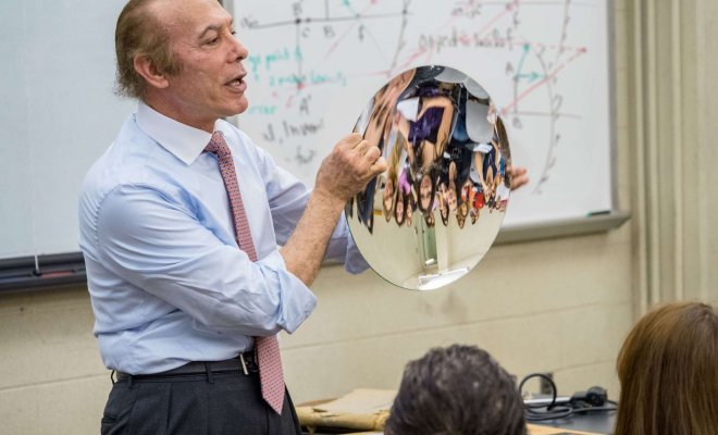 A professor holds a curved mirror in front of a classroom in front of a whiteboard that contains diagrams and formulae for calculating reflections on curved surfaces.
