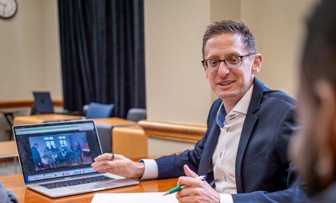 Man is facing the camera explaining while his hand points to his computer.