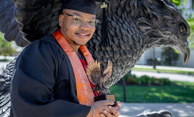 Emmanuel Ozias poses in cap and gown in front of the Red Hawk statute holding a trophy shaped like a red hawk.