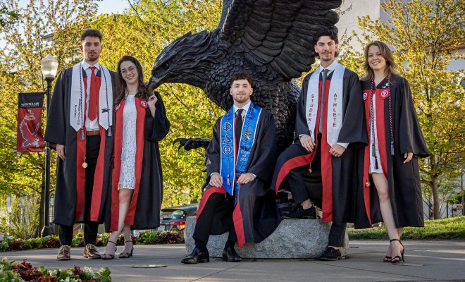 The Povolo quintuplets in graduation gowns stand in front of a large Red Hawk statue.