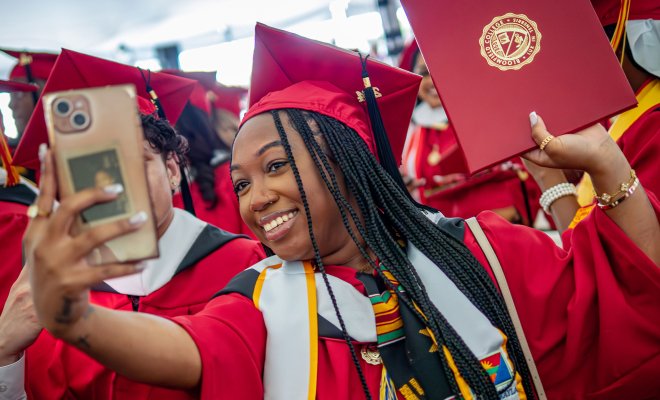 Woman is taking a selfie of herself in a red cap and gown at commencement.