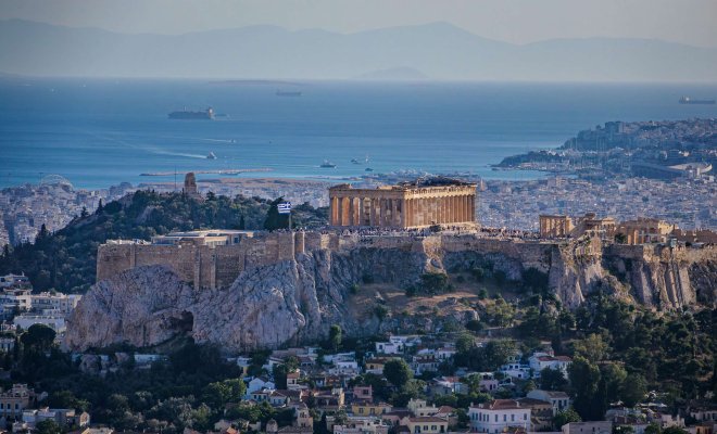 View of the Acropolis in Athens, Greece featuring the Parthenon surrounded by city buildings and overlooking the blue Aegean Sea.