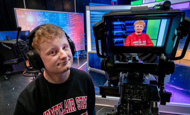 Brian Scott operates a professional television camera in a studio, wearing headphones and a Montclair State University sweatshirt. The camera’s monitor displays his brother, Craig Scott, who is seated on set and visible on the viewfinder, also wearing Montclair apparel.