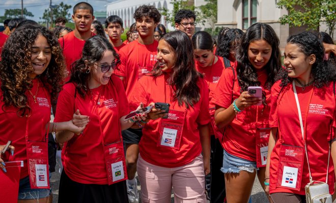 A group of students in red t-shirts and lanyards walk together in a large group on college campus.