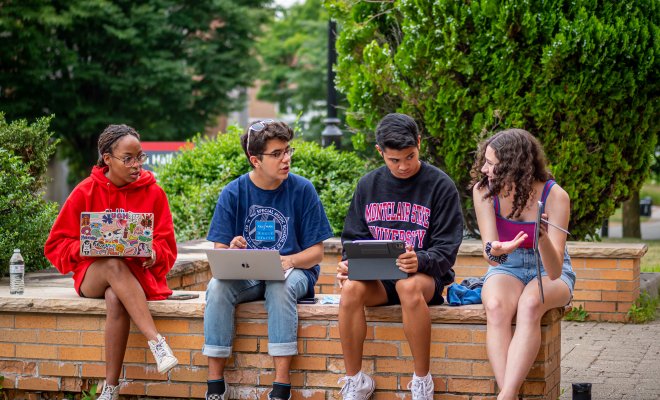 Four students with laptops talk while sitting on a a low wall.