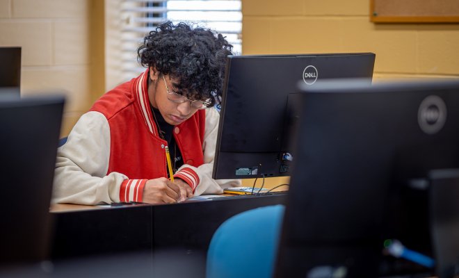 Man in a red letter jacket in front of a computer writing down information.