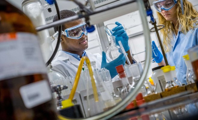 Two students wearing protective eyewear, coats and gloves working together mixing chemicals in a lab.