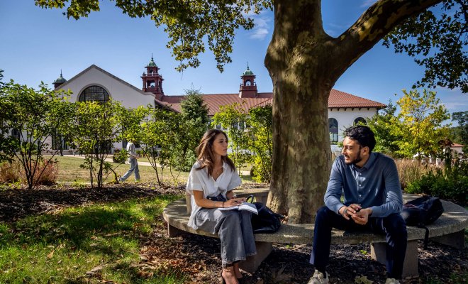 Two students conversing on a bench under tree shade on Montclair campus.