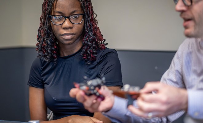 A professor demonstrates a small robot constructed from household items to a student seated nearby.