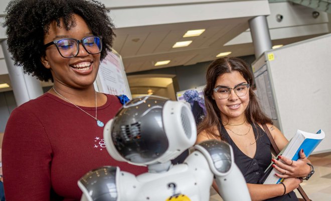 Two young women smiling at symposium as they check out a presentation on robotics.