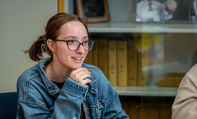 A smiling student sits in a classroom with an open book on the table in front of them.
