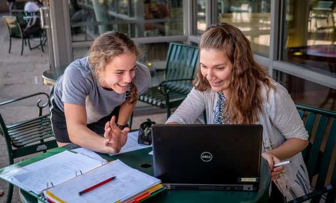 Two female students are seen conversating over a laptop at a table.