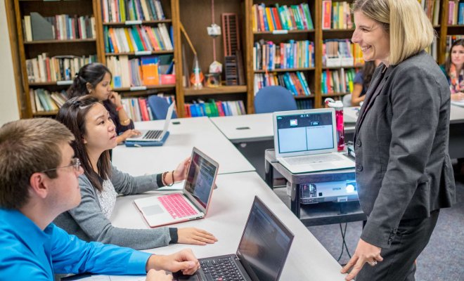 A teacher stands in the middle of a ring of desks talking to two students using laptops. Behind them is a wall of bookshelves.