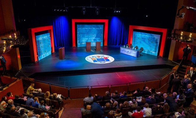 Audience members gather in a theater before a stage set for a debate with podiums, three red archways and the seal of the State of New Jersey on the stage floor.