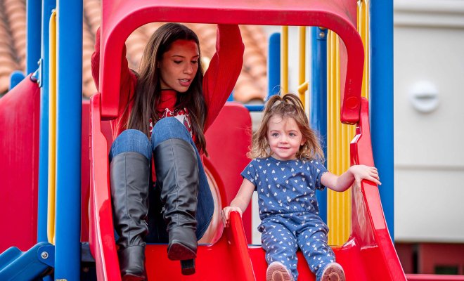 Two people, an adult and a child, are sitting together on a bright red playground slide. The adult wears tall black boots and jeans, while the child is dressed in a blue outfit with small heart patterns.