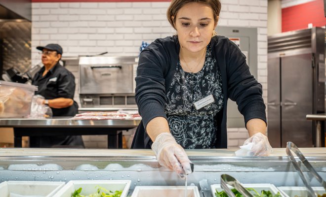 Woman in front of a bar of food with gloves on reaching in.