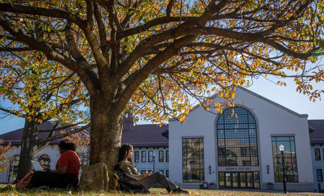 Two students sit under a tree with Cole Hall in the background.