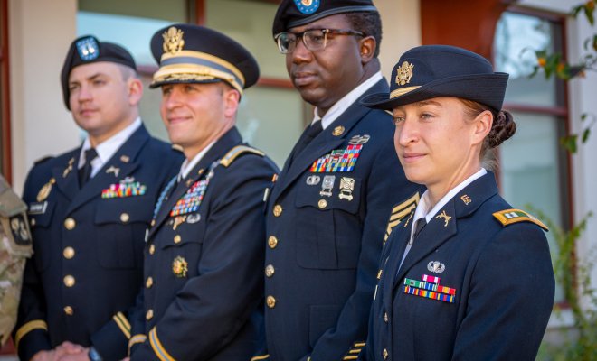 Four uniformed military personnel stand in a row outdoors, wearing formal dress uniforms with caps, medals and rank insignia.