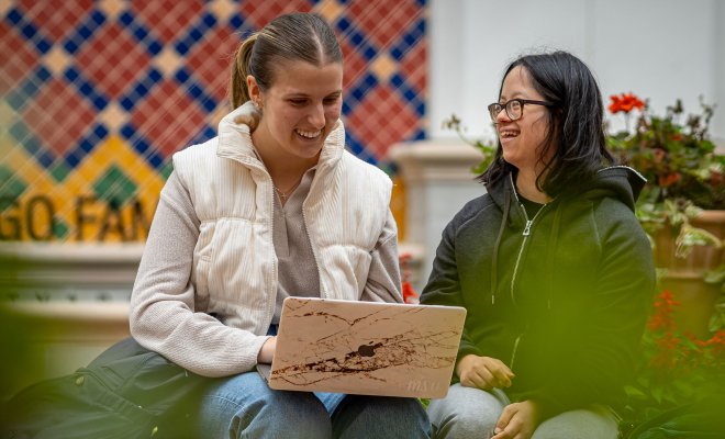 Two students are sitting in an outdoor courtyard, smiling and working on a laptop.