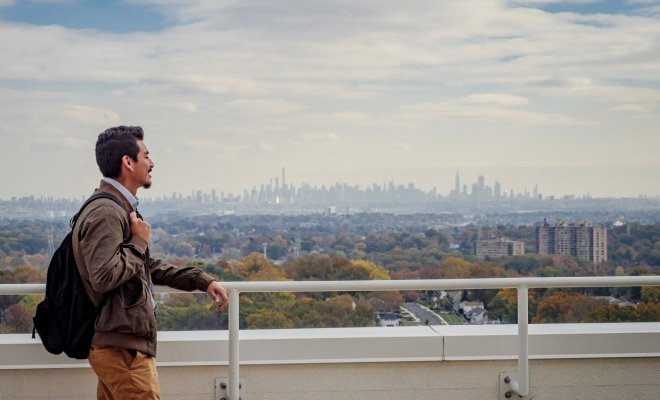 A student stands on a balcony looking out at the Manhattan skyline in the distance.