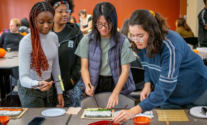 A group of students stands around a table making sushi using bamboo mats and hands, picking up fillings with chopsticks. Various sushi ingredients such as cucumber and carrot sticks, egg slices as well as bowls and plates are arranged on the table. The activity takes place in a classroom with others participating in the background.