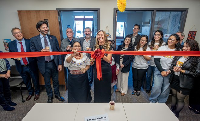 A smiling woman in a black dress cuts a ceremonial red ribbon in a classroom as spectators look on.
