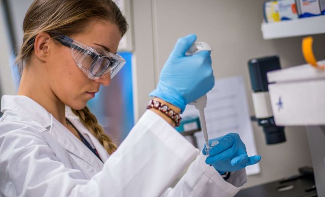Woman student in lab coat with safety glasses and gloves uses a pipette to place a sample into a vial.