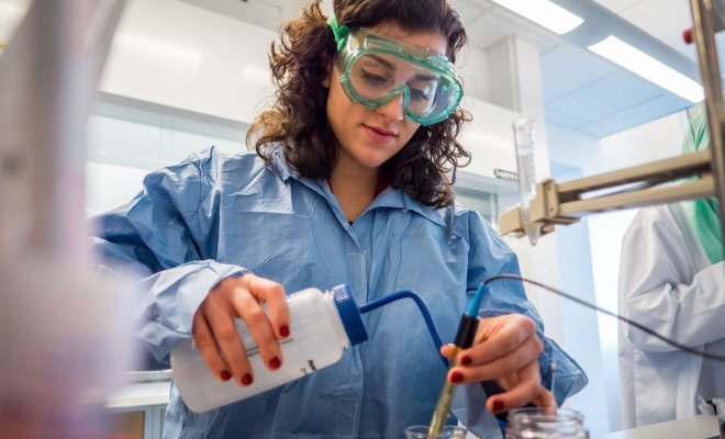 A student in a lab coat with safety goggles carefully mixes chemicals in a beaker.