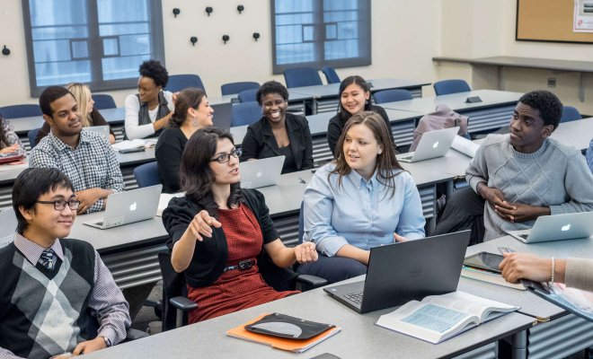 In a classroom with 11 students, a student in a red dress speaks while others listen or interact with their neighbors.
