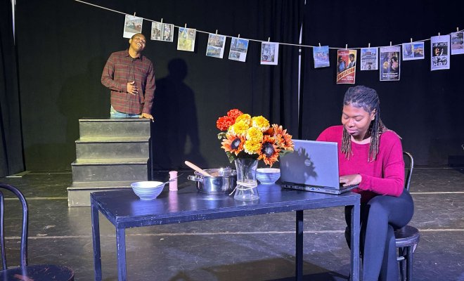 Student working on laptop, seated on a table in stage set.