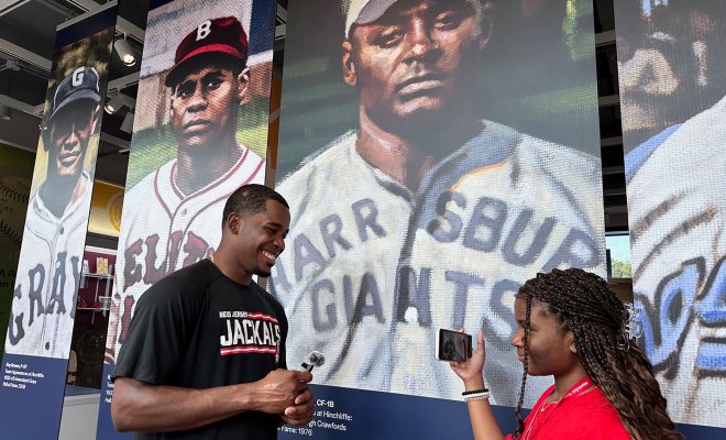 A young reporter interviews a New Jersey Jackals baseball player in front of a display featuring historical baseball players at the Hinchliffe Stadium exhibit.