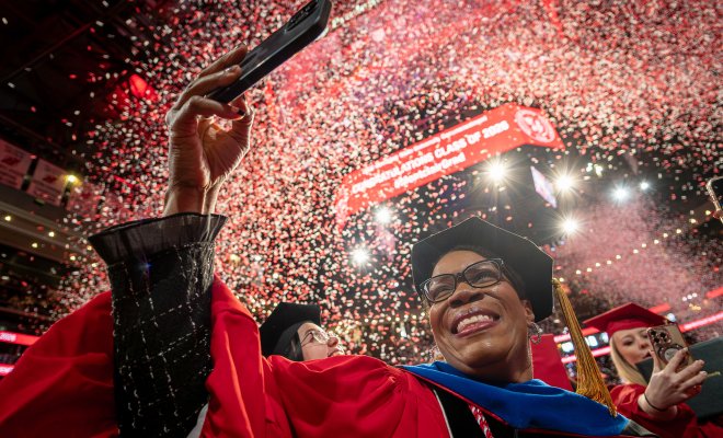 A doctoral graduate in a red gown and black doctoral cap smiles and raises a phone to capture the moment as red and white confetti rains down inside Prudential Center.