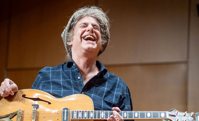 Pat Metheny laughs on stage as he holds a guitar in his lap.