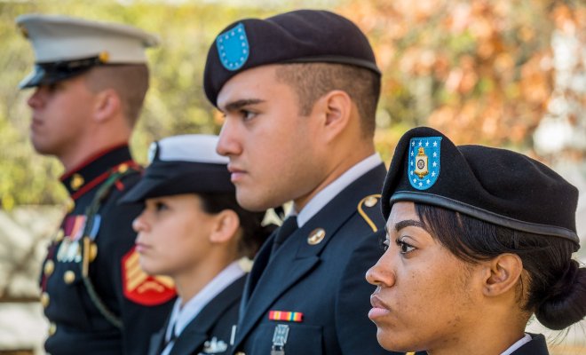 Four U.S. service members stand in a row outdoors, dressed in different military uniforms and caps or berets.