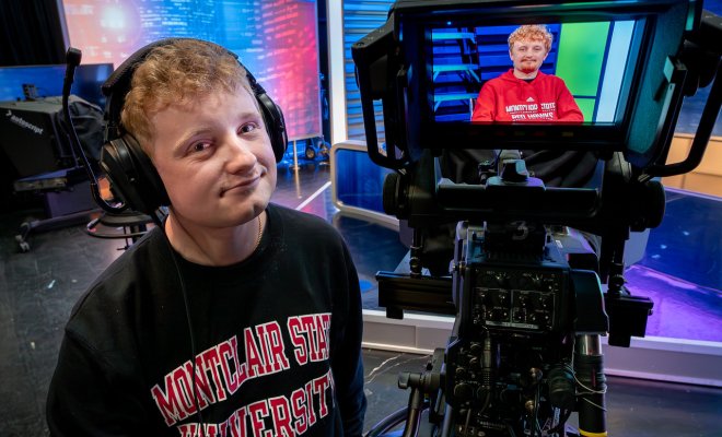 Student in a “Montclair State University” sweatshirt operates a TV studio camera, while another student in a red sweatshirt appears on the monitor in a broadcast set.