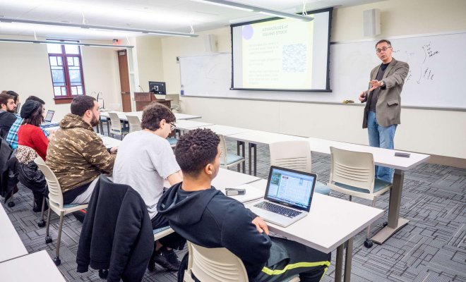 Professor speaking to students in a small classroom with projection screens behind