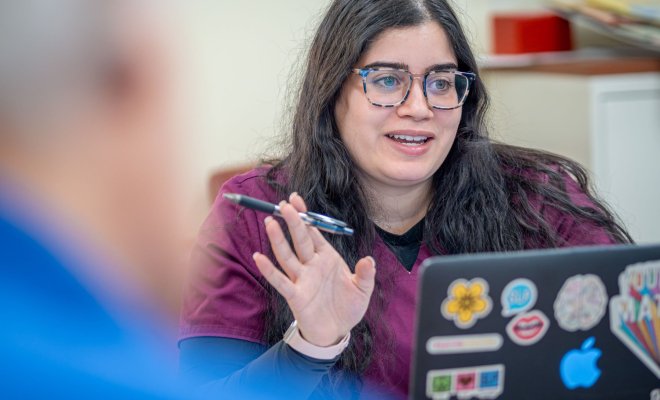 A woman with pen in hand converses with her colleagues while seated behind a laptop adorned with stickers that have messages of positivity.