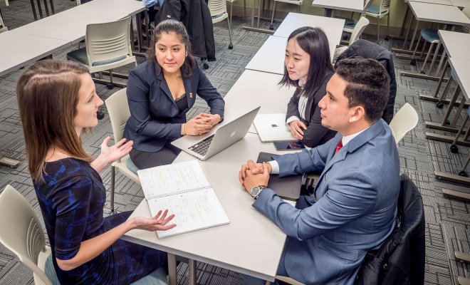 Four students sit at a table with notebooks and laptops open between them.