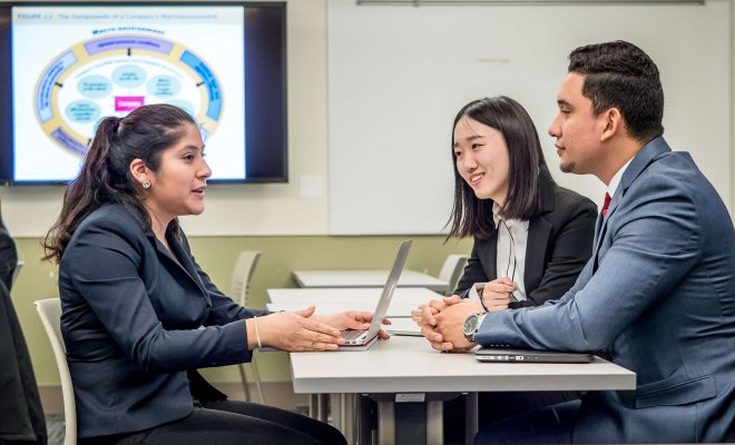 One student with a laptop sits on one side of a table while two others sit across from her, as if in an interview.