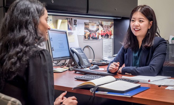 A woman behind a desk in an office speaks with a peer sitting across the desk.
