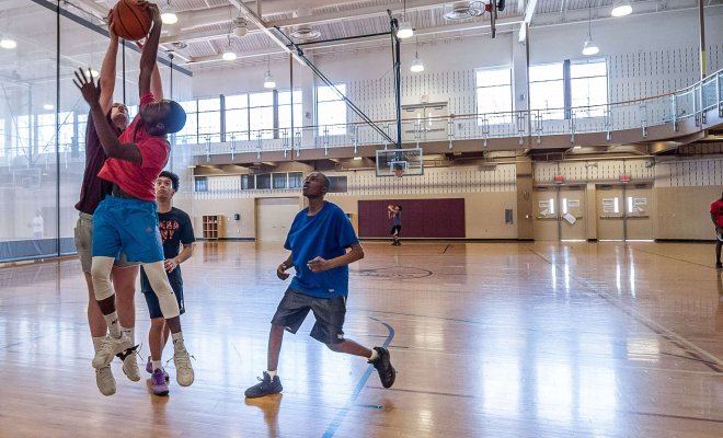One student on a basketball court goes for a lay-up while another student jumps into the air to block the shot.