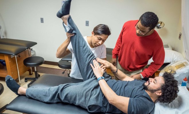 One student laying on a table describes how he's feeling to a trainer as the trainer stretches the student's hamstring. Another student looks on, observing the trainer.