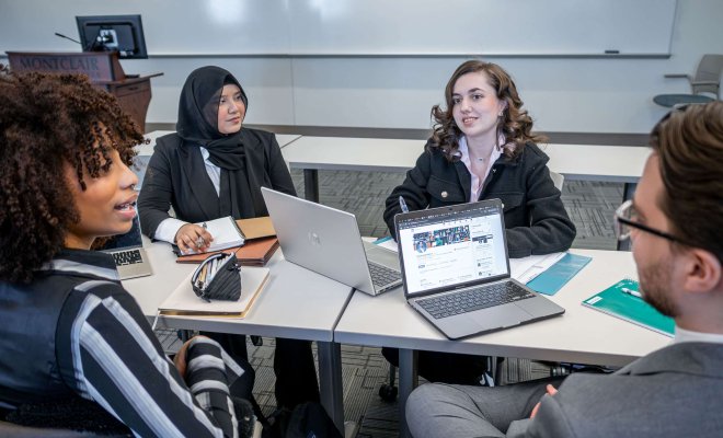 Students in conversation, sitting around table with laptops and notebooks.