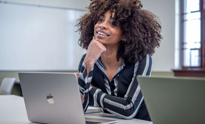 Woman student with laptop sits at table in classroom resting her chin on her hand and smiling.