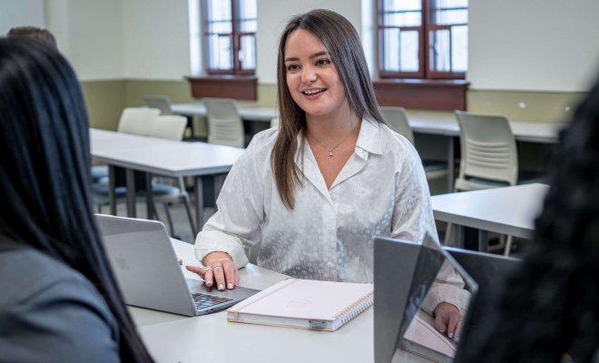 Woman student with laptop and notebook chatting with fellow students off-camera.