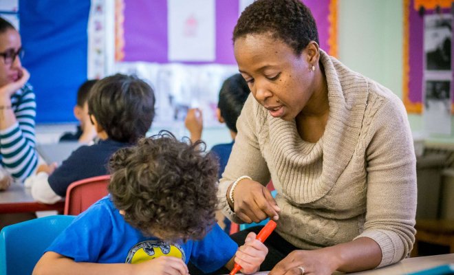 A teacher in a classroom helps a young elementary school student with a coloring assignment.