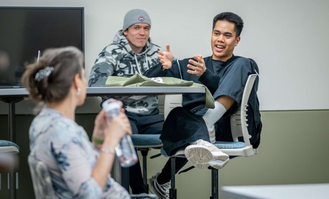 A student seated at a high table with barstools speaks while gesturing with his hands, his professor out of focus in the foreground listening.
